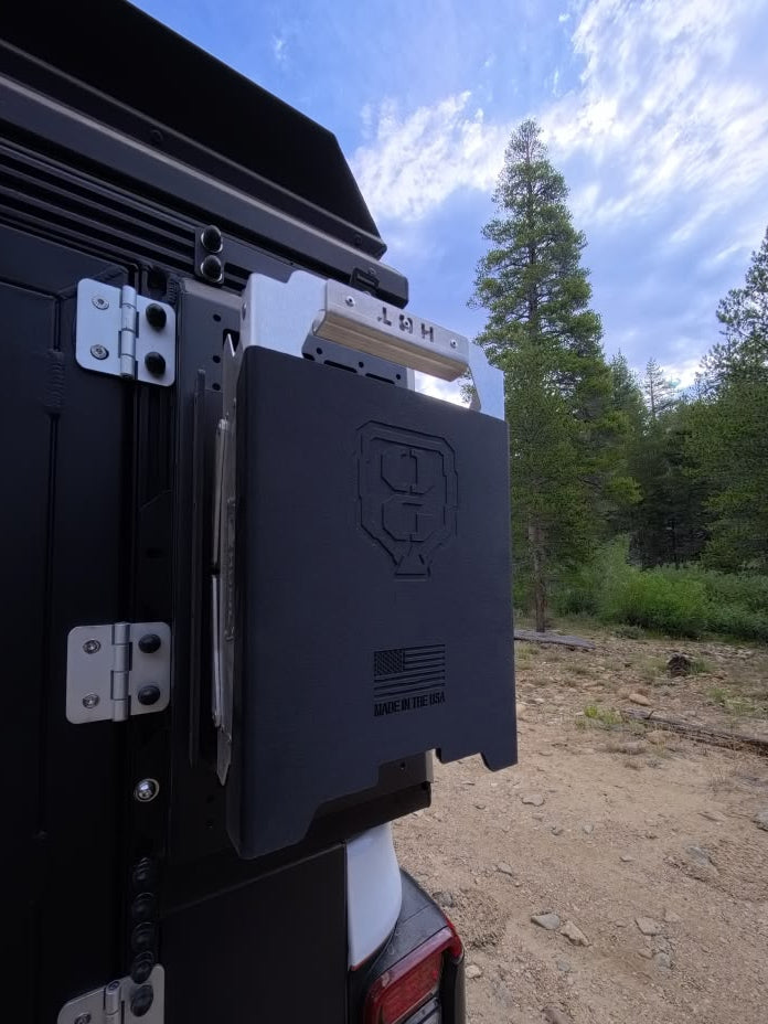 A black metal outdoor utility box or mounting bracket attached to the side of a black vehicle, photographed outdoors with trees and dirt ground in the background.