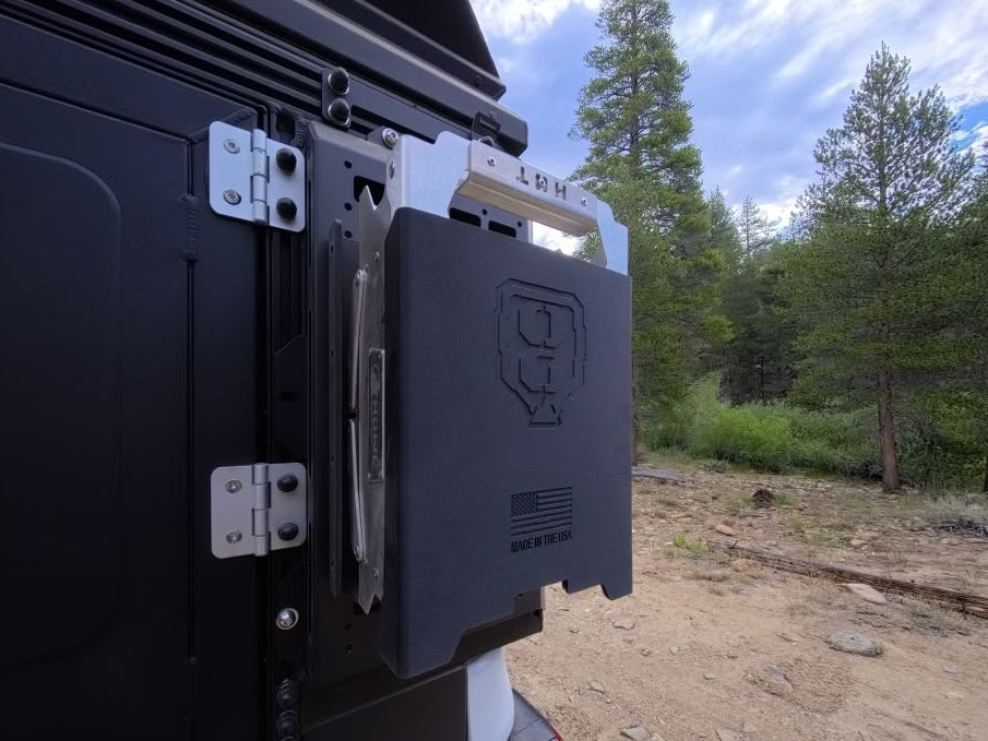 A close-up outdoor shot of a black off-road or camping storage unit mounted to the side of a vehicle, with a forest and dirt road visible in the background. 