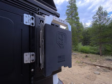 A close-up outdoor shot of a black off-road or camping storage unit mounted to the side of a vehicle, with a forest and dirt road visible in the background. 