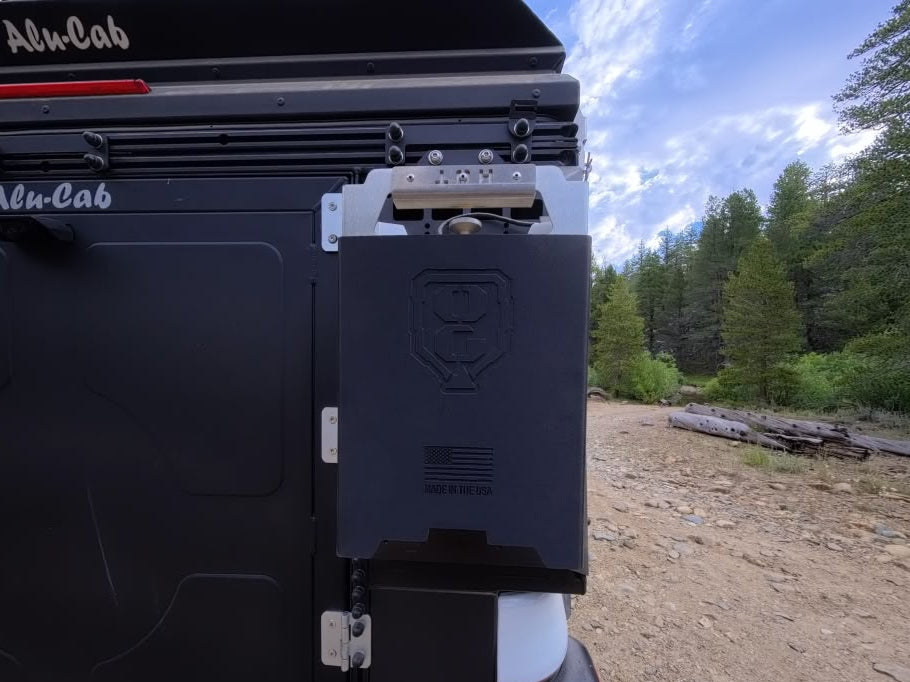 Close-up of the rear side of a black Alu-Cab camper or trailer, showing a mounted black storage box with a silver latch, outdoors on a dirt path with trees and sky in the background.